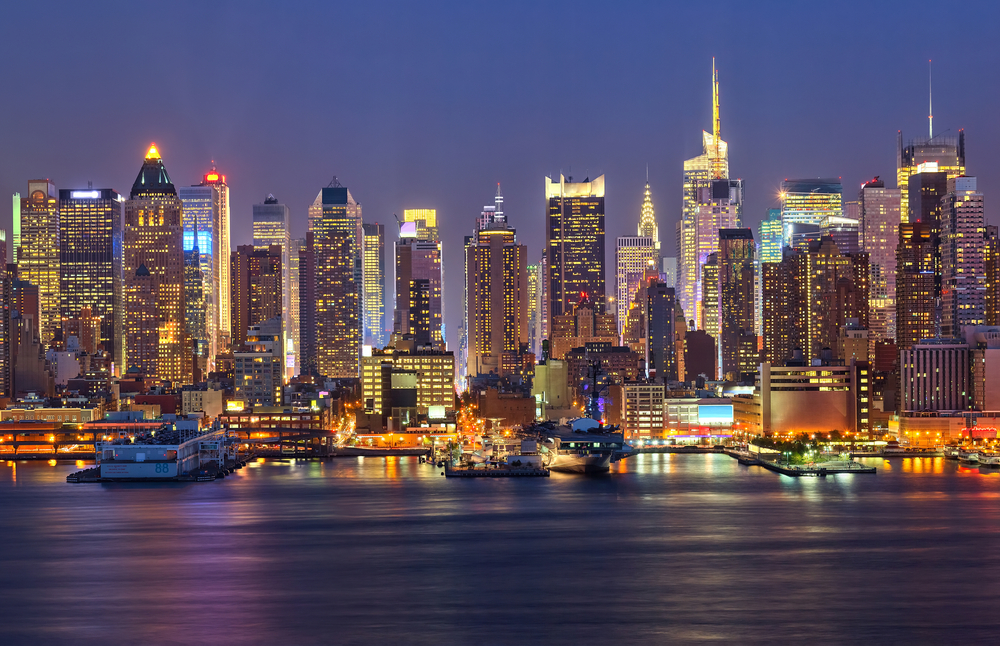 A city skyline view of Manhattan filled with very tall buildings, and water in the foreground at night.