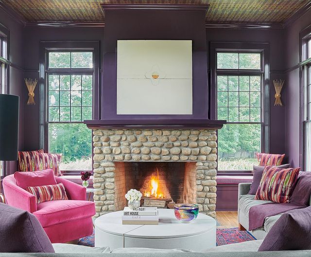 A room with a dark shade of purple painted on the walls, a fireplace in the middle and various furniture in the space.
