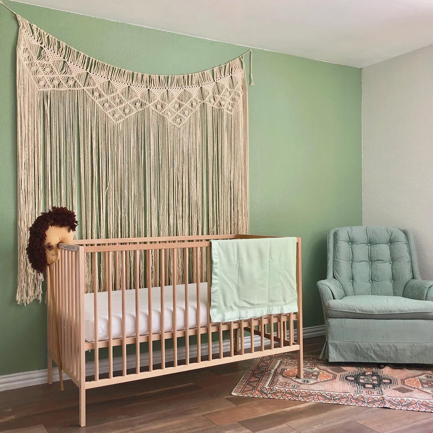 Sage green nursery with large macrame wall hanging and rocking chair. Photo by Instagram user @_sincerelyus