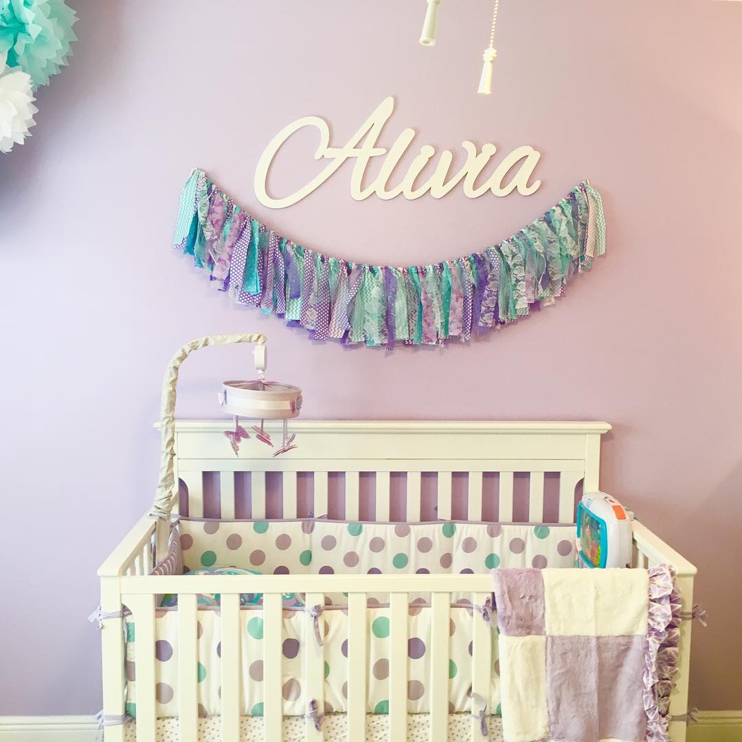 A lavender nursery with a bright garland over the crib and wall art with the baby's name, Alivia, over the crib. Photo by Instagram user @moonsnailcreations.