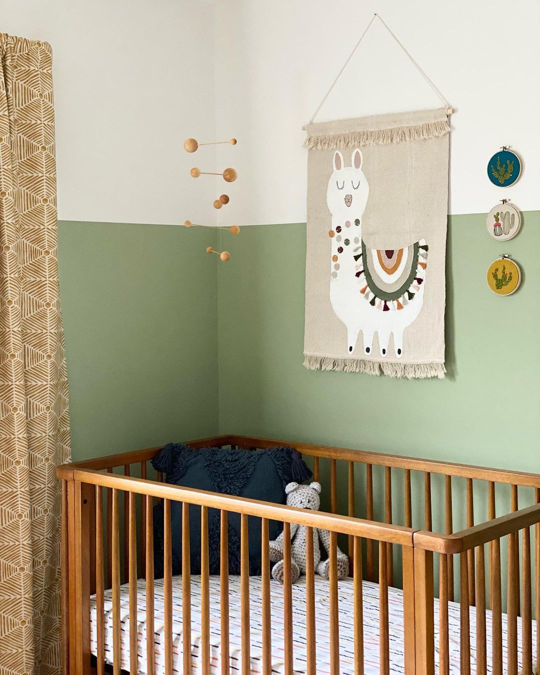 A nursery with the wall painted green halfway up, then white above that point. There is an alpaca tapestry hanging above a wooden crib. Photo by Instagram user @halfacrehomedesign.