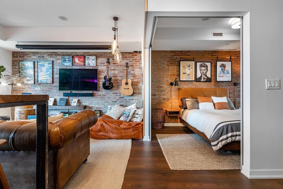 A decorated studio apartment living room and bedroom area with leather furniture, a TV on the wall with art and guitars, and a made bed. Photo by Instagram user @mandustrial_revolution
