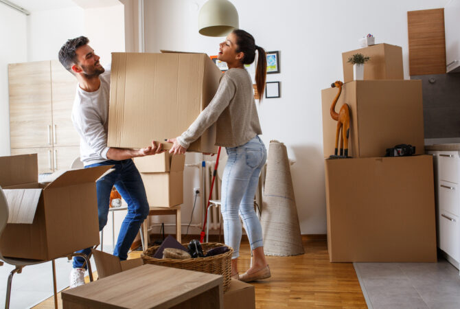 Two people carry a box through a packed-up room.