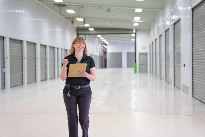 A smiling blonde woman walks through a drive-in storage facility while holding a clipboard.