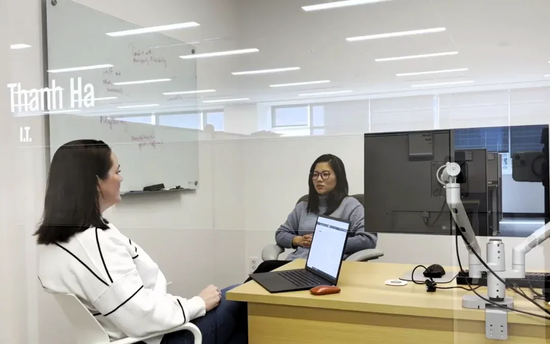 Two women sitting beside a desk in a glass office talking