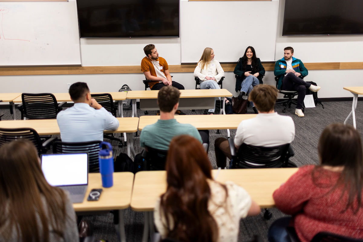 People seated at desks with four speakers also seated chatting at the front of the classroom