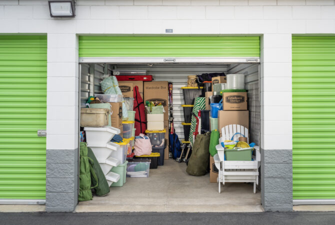 Extra Space Storage unit with green garage door open and filled with storage bins, cardboard boxes, and furniture