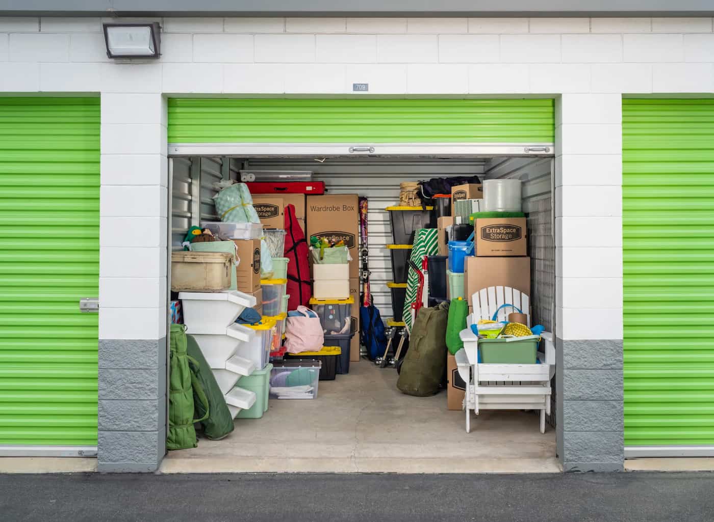Extra Space Storage unit with green garage door open and filled with storage bins, cardboard boxes, and furniture
