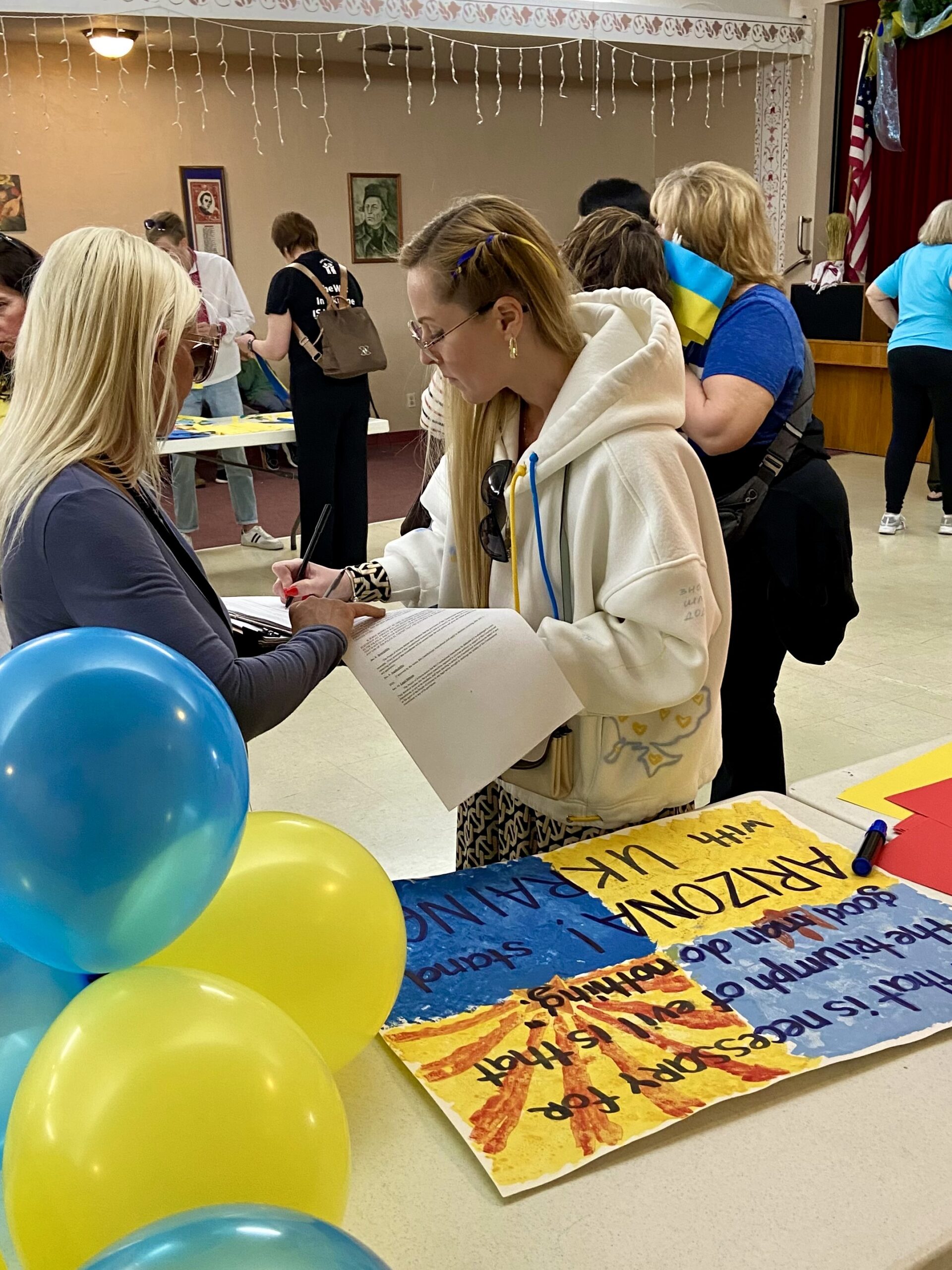 Anastasia Krecklow inside auditorium with yellow and blue balloons and hand-painted signs while signing documents on a clipboard for the Winter Survival Essentials Drive for Ukraine with the Central Park Angels
