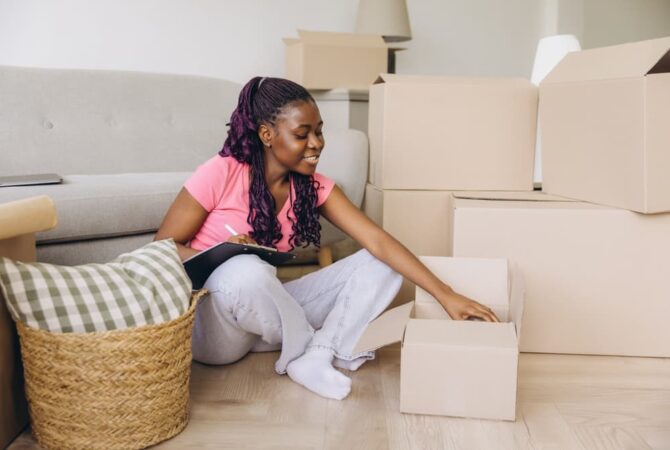 Photo shows a young, Black woman moving into her apartment with a couch behind her, cardboard boxes to the right, and a basket with a pillow to the left. The woman sits on the floor unpacking boxes.