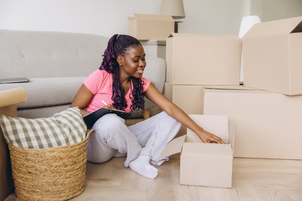Photo shows a young, Black woman moving into her apartment with a couch behind her, cardboard boxes to the right, and a basket with a pillow to the left. The woman sits on the floor unpacking boxes.