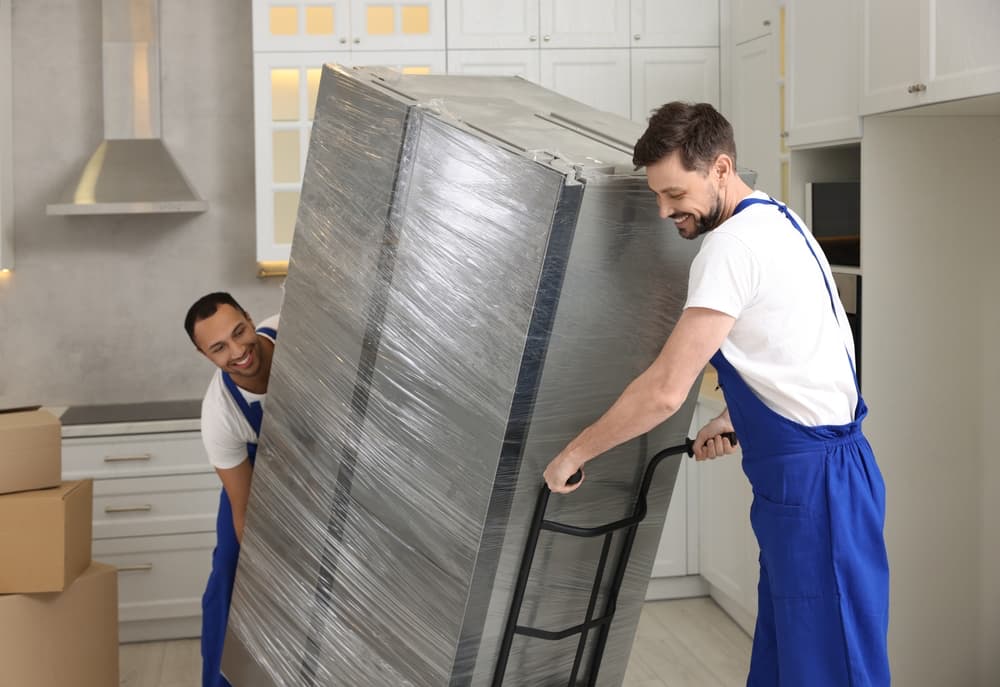 Two people moving a wrapped refrigerator on a dolly